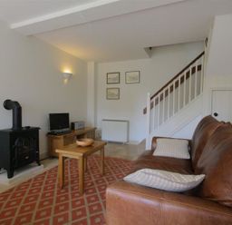 A living room with a brown leather sofa a wooden coffee table a black stove and a wooden media cabinet with a TV at Noel Cottage in Chipping Campden