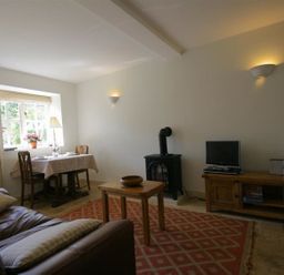 A living room with a brown leather sofa, wooden table, dining table by a window, wood stove, TV on wooden cabinet at Noel Cottage in Chipping Campden
