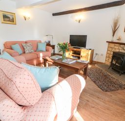 A living room with pink sofas and blue cushions a wooden coffee table with flowers a TV on a wooden stand and a stone fireplace at Cowfair Cottage in Chipping Campden
