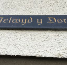 A black rectangular sign with gold Gothic lettering on a white textured wall at Aelwyd Y Don in Tresaith