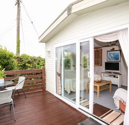 A patio with two chairs and a small table next to a cabin with sliding glass doors revealing a living area with a sofa a coffee table and a television inside at Bwthyn Y Bae Lodge Church Bay near Llanfaethlu