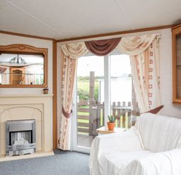A living room with a fireplace a white covered sofa glass cabinet and glass door with curtains at Bwthyn Y Bae Lodge in Church Bay near Llanfaethlu