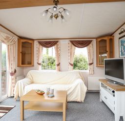 A living room with a sofa covered in a light throw a wooden coffee table a television on a white stand and curtains on windows at Bwthyn Y Bae Lodge in Church Bay near Llanfaethlu