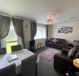 A living room with brown sofas and purple cushions a dining table with chairs a small tv and windows with curtains at Lands End in Liskeard near St Cleer