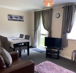 A living room with a dining table and chairs a television on a stand and a brown sofa at Lands End in Liskeard near St Cleer