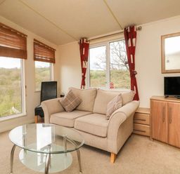 A living room with a beige sofa, glass coffee table, black chair, and wooden cabinet with a TV at Exe Valley Lodge in Bampton Devon