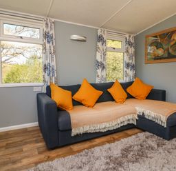 A sitting room with a dark blue sofa with orange cushions and beige throws next to windows with floral curtains at Valley Lodge in Bampton Devon
