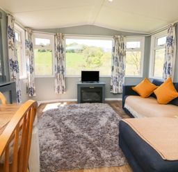 A living room with a wooden dining table and chairs a gray carpet and blue sofas with orange cushions at Valley Lodge in Bampton Devon