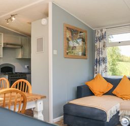 A dining table with wooden chairs and a kitchen area with stove beside a living area with a couch and orange cushions at Valley Lodge in Bampton Devon