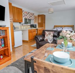 A kitchen dining and living area with wooden cabinets a dining table set with bowls and wine glasses a brown leather sofa and a bed in the background at The Pool House in Crediton