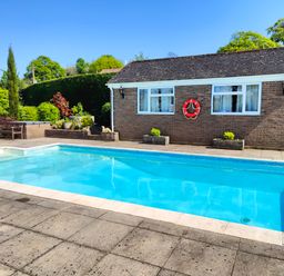 An outdoor swimming pool with a bench and greenery next to a brick building at The Pool House in Crediton
