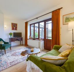 A living room with a green sofa, wooden coffee table, armchair, desk with TV, and large window with orange curtains at Little Barton in Stogumber near Watchet
