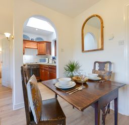 A dining area with a wooden table set for two next to an archway leading to a kitchen with wooden cabinets and a bedroom visible through an open door at Little Barton in Stogumber near Watchet