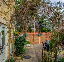 A garden area with a stone pathway a metal arch with climbing plants outdoor table and chairs near a brick wall at Jasmine Cottage in Mickleton Gloucestershire
