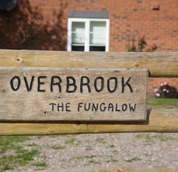 A wooden sign with the text Overbrook The Fungalow on a wooden fence with a brick house in the background at Overbrook in Salwarpe near Droitwich Spa