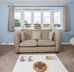A living room with a beige sofa pillows a white coffee table with books and a wooden game set blue walls and a bay window with curtains at Overbrook in Salwarpe near Droitwich Spa