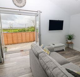 A living room with a gray sofa cushions a wall mounted TV and an open door leading to a patio at Caban Y Mor in Amroth near Saundersfoot