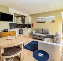 A living room with two gray sofas a wooden dining table a small blue side table a wall mounted television and a kitchenette at Chalet 126a in Cromer