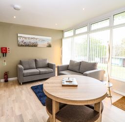 A living room with two gray sofas a round wooden table and large windows with vertical blinds at Chalet 126a in Cromer