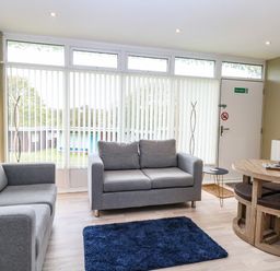 A living room with two gray sofas a blue rug a round wooden dining table and chairs near a door with vertical blinds at Chalet 126a in Cromer