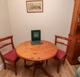 A round wooden table with two wooden chairs with red cushions and a wooden cabinet with books in a room at Rose Cottage in Akeld near Wooler