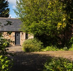 A stone building with a door and window surrounded by trees and bushes at Bramble Cottage in Akeld near Wooler