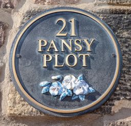 A round plaque with number 21 and Pansy Plot on a stone wall with flower decoration at Pansy Plot Akeld near Wooler