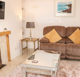 A living room with a brown sofa, yellow cushions, a glass coffee table, a side table with a lamp, a fireplace, and a wall-mounted TV at Lane Cottage in Ballycullane County Wexford