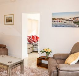 A living room with two brown armchairs a glass coffee table a small wooden side table with flowers a wall painting of boats and a view into another room with a white metal bed and red pillows at Lane Cottage in Ballycullane County Wexford