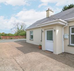 An exterior view of a beige house with a white door a mailbox and windows with vertical blinds surrounded by trees and a wooden fence at Lane Cottage in Ballycullane County Wexford
