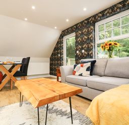 A living room with a gray sofa a wooden coffee table a dining table with chairs and a window with flowers at Robins Nest at The Shire Inn Killivose near Camborne