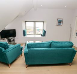 A living room with teal sofa and armchair a glass table and TV near a window at 21 The Rest in Porthcawl