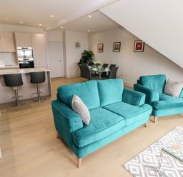 An open plan living room with teal sofas a glass coffee table a dining table with chairs and a kitchen area with stools at 21 The Rest in Porthcawl