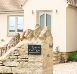 A stone wall with a sign reading Tawny Cottage outside a house with beige walls and grey framed windows at Tawny Cottage in Fulbrook near Burford