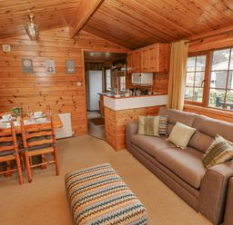 A wooden cabin interior with a dining table set for four a gray sofa with pillows a patterned ottoman and a kitchen area at Heritage Park Stepaside near Kilgetty