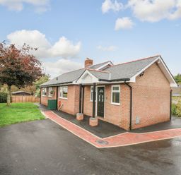 A single story brick house with a black door and windows next to a paved driveway and a lawn with a tree at Ger Yr Afon Cottage in St Harmon near Rhayader