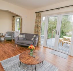 A living room with a round wooden coffee table with flowers on it and grey sofas near glass doors opening to a deck with a picnic table at Ger Yr Afon Cottage in St Harmon near Rhayader