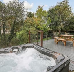 An outdoor deck with a bubbling hot tub and a wooden picnic table with benches surrounded by trees at Ger Yr Afon Cottage St Harmon near Rhayader