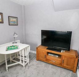 A small room with a television on a wooden stand and a white table with a lamp and a board game at The Old Bakery in Milton Tenby