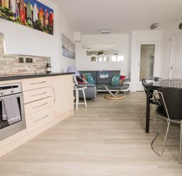 A kitchen and living area with oven and cabinets sofa and glass coffee table and dining table with chairs at Fistral Drift in Newquay