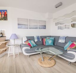 A living room with a curved sofa with colorful cushions a glass coffee table a side table with a lamp and wall art at Fistral Drift in Newquay