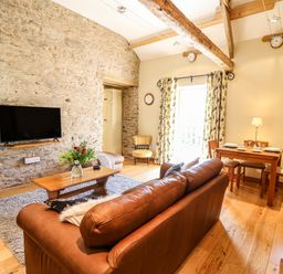 A living room with a brown leather sofa a wooden coffee table flowers on the table a wall mounted tv and a dining table set for four at Golwg Las in Pumsaint near Lampeter
