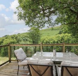 An outdoor patio with a glass table and four cushioned chairs overlooking green trees and hills at Nant Glas in Pumpsaint near Lampeter