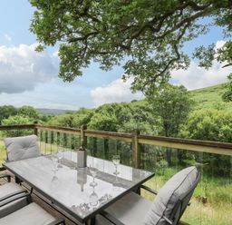 An outdoor patio with a glass table and cushioned chairs overlooking trees and hills at Nant Glas Pumpsaint near Lampeter