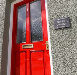 A red front door with the number 1 and letterbox on a textured grey wall with a Kooky Cottage sign in Brecon