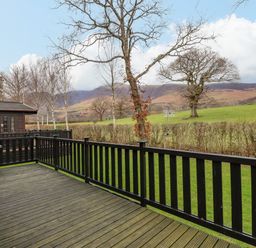 A wooden deck with black railings overlooking a grassy field with trees and hills at The Fold Number 28 Burnside Park near Keswick