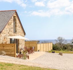 A stone house with a wooden fence patio furniture and a view of trees and the sea at Apple Blossom Barn in Gulval near Penzance