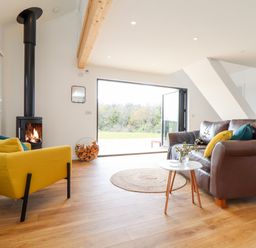A living room with a yellow armchair a brown leather sofa a round rug a wood burning stove and an open glass door at Apple Blossom Barn in Gulval near Penzance