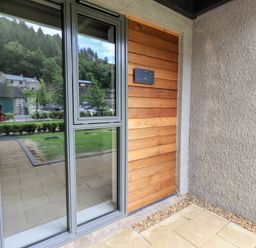 The entrance to a property with a wooden door numbered 24 and large glass windows reflecting trees and nearby buildings at Rivers Edge in Newby Bridge