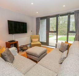 A living room with a large corner sofa and ottoman facing a wall mounted TV and glass doors at Rivers Edge in Newby Bridge